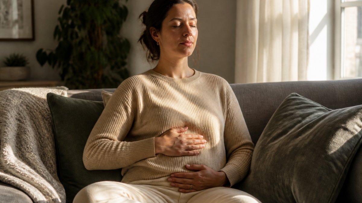 A person sitting comfortably on a couch in a quiet living room, eyes closed, one hand resting on their belly, practicing slow deep breathing in soft afternoon light